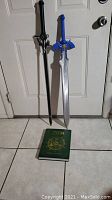 View of both replica swords standing against a white door with the Hyrule Historia book lying on the floor beneath them