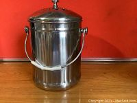 Front view of stainless steel compost pail with handle and lid with a knob, placed on wooden surface against orange background.