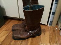 Side profile of both brown leather John Fluevog motorcycle boots standing next to each other on a wood floor.