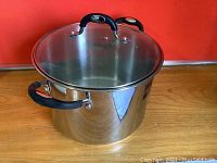 Large stainless steel stock pot with black handles and clear glass lid with handle on top.