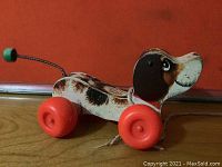 Side and front view of the wooden Little Snoopy pull toy with red wheels and spring tail on brown surface against orange background.