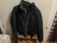 Full view of black Moose Knuckles bomber jacket hanging on door, showing zip front, ribbed sleeve cuffs and hem, front pockets with snap closures.