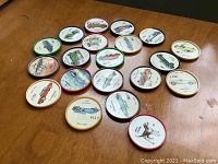 A grouping of twenty round collectible coins/buttons with colored rim edges and paper inserts depicting vintage automobiles and aircraft. Each button is labeled with the model name and year, all well arranged on a wooden surface.