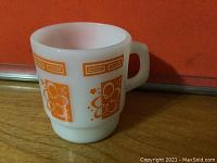 White opaque glass mug with orange psychedelic floral and geometric design on a wooden surface in front of an orange background.
