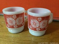 Pair of vintage Fire King mugs with red daisy floral patterns on white glass, showing front view to display design and handles.
