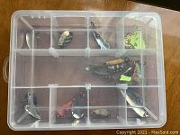Top view of clear plastic tackle box showing several metal and painted fishing lures including spoons and spinners arranged in compartments