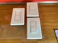 Three white smart switches with dimmer controls arranged on wooden surface in packaging trays, showing front with rocker buttons and indicator lights.