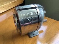 Side view of shiny metal Boston No 8 pencil sharpener mounted on a wooden surface, showing embossed branding and crank handle.