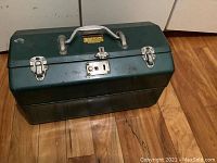 Front view of a green metal tackle/tool box placed on a wooden floor, showing handle, latch locks and condition of the box.