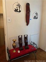 Wide view showing metal bench topped with red cushion, two Buddha figurines, gold vase and portion of wall with clock and two metal art pieces.