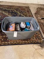 Wide view of gray lidded plastic bin on rug showing contents inside including boxes and cables.