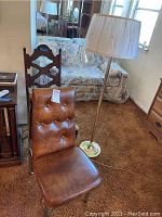Photo shows a brown tufted faux leather chair, wooden carved backrest chair, and brass floor lamp with white pleated lampshade.