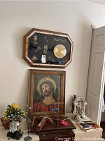 Framed octagonal clock with gold-tone face on black backing with metal decorations, rectangular framed Jesus Christ art, assorted Christian figurines, faux flowers, crucifix, model car, paperweight, and candle lantern on table near a wall.