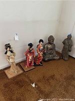Full view showing the three geisha dolls in traditional attire with wooden bases, seated Buddha statue, and standing robed figure statue placed side by side on a brown carpet against a white wall.