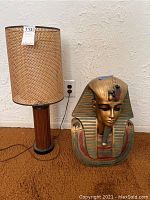 Table lamp with woven cylindrical shade and wooden base next to Egyptian Pharaoh bust statue.