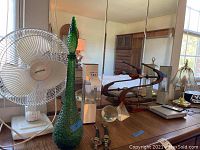 Oscillating fan, green glass bottle, cutters with sheaths, paperweights, lamp with glass shade, and part of DVD player visible on wooden table against mirrored wall.
