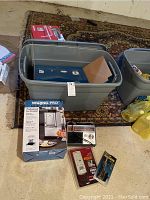 Overview of lot items showing gray lidded storage bin containing various items including a blue box and packing material, Waring Pro ice crusher box, a radio and other small packaged items