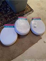 Three white Softy brand toilet seats in original shrink wrap packaging resting on carpet and floor.