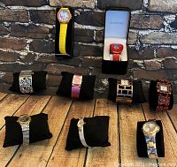 Front view of nine assorted watches placed on black cushions on wooden surface against stone wall background, showcasing a variety of designs and band types.