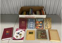 Photo showing assortment of vintage books arranged with several laid out in front of cardboard box containing more books.