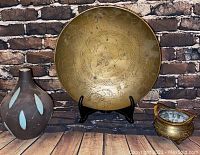 Photo showing the three items: large brass decorative bowl on stand, small brass tripod bowl with handle, and brown ceramic vase with white leaf patterns on wooden surface against brick wall background.