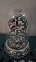 Front view of Mickey Mouse anniversary clock under clear glass dome, showing clock face with Mickey's arms as hands and decorative base with Mickey images.