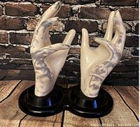 Pair of white ceramic hands with marbled gray pattern mounted on a black plastic base, positioned on wood surface with a brick background.