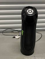 Front view of black cylindrical electric heater showing mesh grille, green side switch, and power cord plugged out.