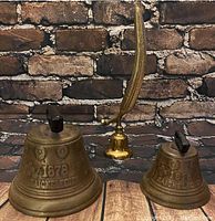 Two Saicelecies metal bells dated 1878 and a tall brass feather-shaped bell in front of brick wall background