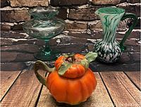 Three vintage glass items on a wooden surface with stone backdrop: two glass vases and one ceramic pumpkin sugar bowl with lid removed.