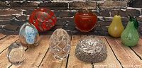 Photo showing seven assorted vintage glass pieces arranged on a wood floor and brick backdrop, including Murano glass fruits (pears, two red/orange fruits), clear cut crystal paperweights and a floral motif glass egg.