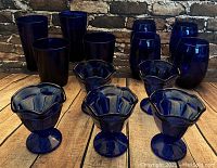 Front view of all glass pieces displayed including tumblers, rocks glasses, and fluted dessert bowls arranged on wooden surface against a brick wall.