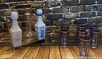 Three various vintage decanters with Asian motifs and metal stand, with six blue and gold decorated shooter glasses on wooden table with brick wall background.