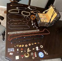 Overview of assorted vintage jewelry spread on dark wooden table including necklaces, rings, pins, pendants, and a jewelry box with compartments.