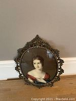 Full frontal view of the oval framed print showing the lady in a red gown surrounded by ornate brass leaf and scroll detailing in the frame.