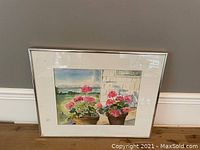 Framed watercolour painting showing two pots with bright pink flowers against a scenic backdrop including water and greenery, hung on a gray wall.