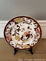 Front view of round ceramic cabinet plate with floral and scrolling designs in brown, yellow, orange, and green on white background, standing on display stand.