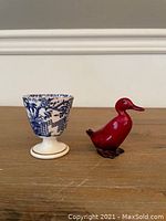 Side-by-side photo showing the blue and white Royal Crown Derby Mikado egg coddler and the red Royal Doulton Flambe duck figurine on a wooden surface.