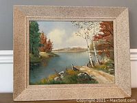 Framed oil painting showing a lakeside scene with autumn foliage and birch trees, signed by the artist in the lower right corner.