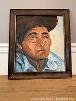 Framed oil on board painting of an elderly person wearing a large hat and light blue clothing, signed lower right.