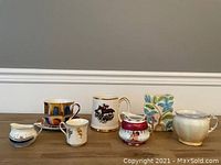 Seven assorted cups and creamers of various designs and sizes shown together on table.