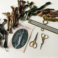 Overview of scissors, sewing tools, embroidery thread, and needlepoint bars arranged on a surface.