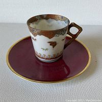 Front view of the demitasse cup on its saucer showing the cup's shape and the maroon saucer with gold rim.