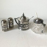 Four items including pair of silver-plated salt and pepper shakers with decorative pierced silver and blue glass liners, silver-plated lidded condiment container with a spoon, and cut glass lidded container with hinged lid and spoon on white background.