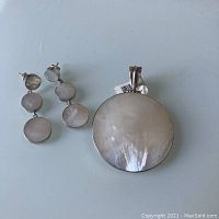 Silver earrings with three circular mother of pearl discs linked vertically next to a large round mother of pearl pendant with silver edges laid flat on a white surface