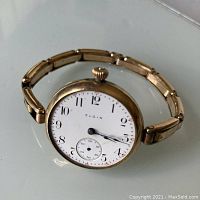 Front view of the gold-tone wristwatch showing white dial with black Arabic numerals and sub-second dial, with blue hands, missing face crystal.