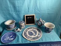 Photo showing the full assortment of seven blue and white ceramic and porcelain items arranged on a blue fabric background with lot number sign.