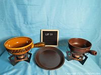 Front view showing two ceramic fondue pots with handles and black metal burner stands, plus one brown ceramic plate in the middle.