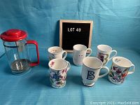 Full lot overview with Bodum coffee press and six assorted floral and lettered coffee mugs on blue background next to sign reading 'LOT 49'