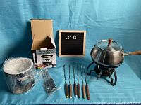 Photo of opened cheese fondue set with pot, lid, black metal stand, burner, and six fondue forks with wooden handles alongside unopened chocolate fondue set in its original box.